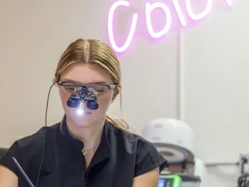 A lab technician wearing magnifying loupes hand-staining a crown under a neon Color sign.