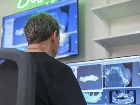 A lab technician at two screens designing a restoration from a 3D scan, under a neon Design sign.